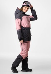 Completo da sci rosa e nero con cappuccio, diverse tasche con cerniera e polsini rinforzati. Indossato con occhiali e stivali neri.
