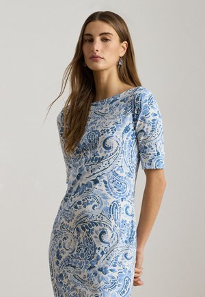 Vrouw met lang bruin haar, gekleed in een nauwsluitende witte jurk met blauwe bloem- en paisleypatronen, en bijpassende blauwe oorbellen.