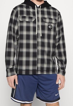 Mężczyzna ubrany w czarno-szaro kraciastą, zapinaną na guziki bluzę z kapturem i kieszeniami z przodu oraz niebieskie sportowe spodenki Nike z białymi paskami.