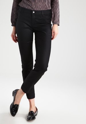 Femme portant un pantalon noir ajusté, un chemisier à manches longues à motif et des mocassins noirs brillants, debout, jambes croisées.