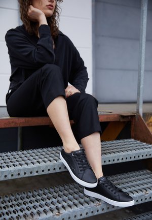 Persona con ropa negra sentada en escaleras de metal, apoyando la cabeza en la mano, usando zapatillas negras con suelas blancas.