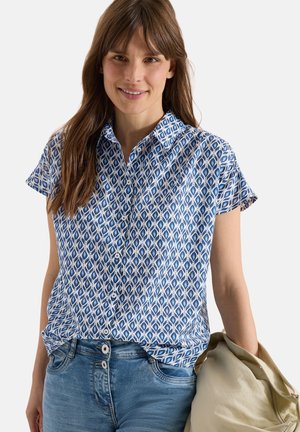 Kobieta uśmiechająca się, ubrana w niebiesko-białą koszulę na krótki rękaw z guzikami oraz jasnoniebieskie jeansy, trzymająca beżową kurtkę w prawej ręce.