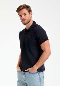 Marineblå polo t-shirt med et struktureret design, korte ærmer og krave med lynlås. Har et lille logo på brystet og sideskår for komfort.