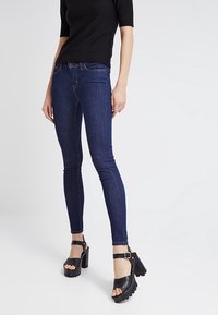 Jeans ajustados de color azul oscuro con cintura alta y costuras en contraste, combinados con sandalias negras de tacón grueso y con correas alrededor del tobillo.