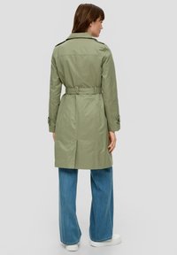 Oliwkowy trencz z paskiem w talii, kołnierzem i zapinanymi mankietami, w połączeniu z jasnoniebieskimi szerokimi spodniami dżinsowymi i białymi sneakersami.