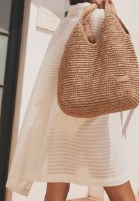 Spleciona torba hobo z trawy w jasnobrązowym kolorze, o pojemnym okrągłym kształcie oraz podwójnych uchwytach, z małą naszywką z logo, na tle paskowanej białej tkaniny.