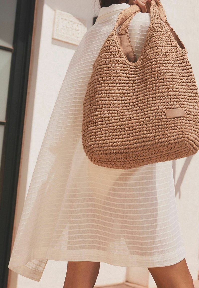 Spleciona torba hobo z trawy w jasnobrązowym kolorze, o pojemnym okrągłym kształcie oraz podwójnych uchwytach, z małą naszywką z logo, na tle paskowanej białej tkaniny.