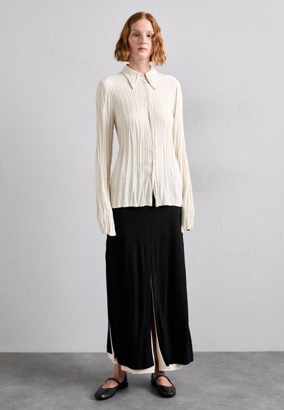 Off-white, pleated long-sleeve blouse with button closure, paired with a black, ankle-length skirt featuring a front split and slip detail.