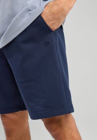 Navy blue cotton shorts featuring side pockets and a small logo on the left hip. The fabric has a smooth texture with stitched hems.