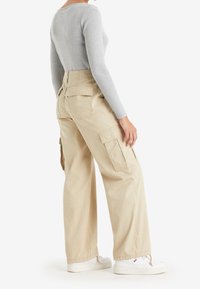 Osoba oblečená v béžových širokých cargo kalhotách, šedém žebrovaném topu s dlouhým rukávem a bílých teniskách, stojící proti jednoduchému bílému pozadí.