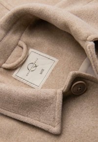 Cappotto di lana beige chiaro con colletto a revers, dotato di un'etichetta cucita e un dettaglio con bottone sul davanti. Tessuto morbido e testurizzato.