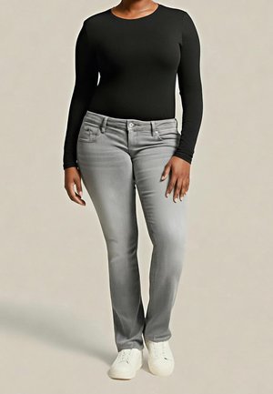 Person iført en sort langærmet top, lysegrå jeans og hvide sneakers, stående mod en ensfarvet lys baggrund.