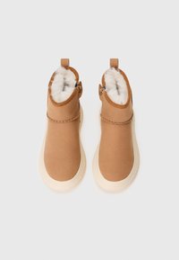 Botas de tobillo marrones con un suave forro de piel blanca, acentos cosidos alrededor de la parte superior y una suela de goma de color crema claro. Incluyen cremalleras laterales.