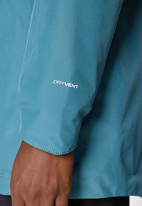 Blå vanntett jakke med en glatt tekstur og en tettsittende design. Har et "DRYVENT"-logo på ermet.