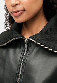 Μαύρο δερμάτινο μπουφάν με φαρδιά ριχτή κολάρα, ασημί φερμουάρ και λεία υφή. Κοντινό πλάνο της κολάρας και της περιοχής του φερμουάρ.