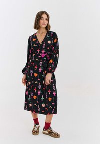 Czarna sukienka kopertowa z kolorowym wzorem owoców i motywów, z długimi rękawami i wiązaniem w talii. Noszona z wzorzystymi trampkami i czerwonymi skarpetkami.