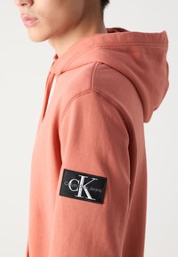 Felpa color rosa con una patch nera che presenta un marchio bianco. Tessuto morbido, con dettagli di cuciture visibili e cappuccio con cordini.