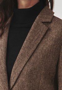 Ruskea teksturoitu bleiseri, jossa on lovettu kaulus, kerrospukeutuneena mustan ribbineuleisen poolopaidan päälle. Keskittyminen kankaan yksityiskohtiin ja värikontrastiin.