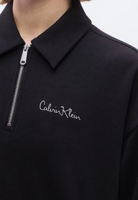Polo nera con zip realizzata in tessuto strutturato con un logo ricamato in bianco sul lato sinistro. Presenta un colletto classico e dettagli in metallo argentato.