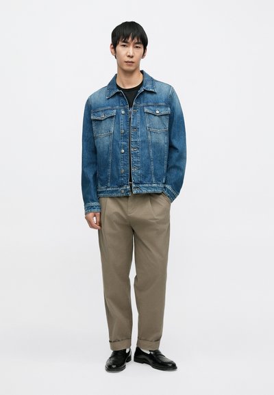 Veste en denim bleu clair avec fermeture à boutons, deux poches poitrine et col classique. Associée à un pantalon beige et des chaussures noires.