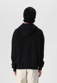 Hanorac negru cu glugă, cu o croială lejeră, manșoane ribolite și o culoare uniformă. Materialul pare moale, cu o textură netedă.