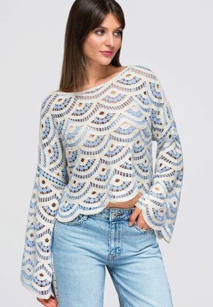 Kvinde iført en hvid og blå hæklelangærmet top med bølgekant og lyseblå jeans, der poserer med den ene hånd i lommen.