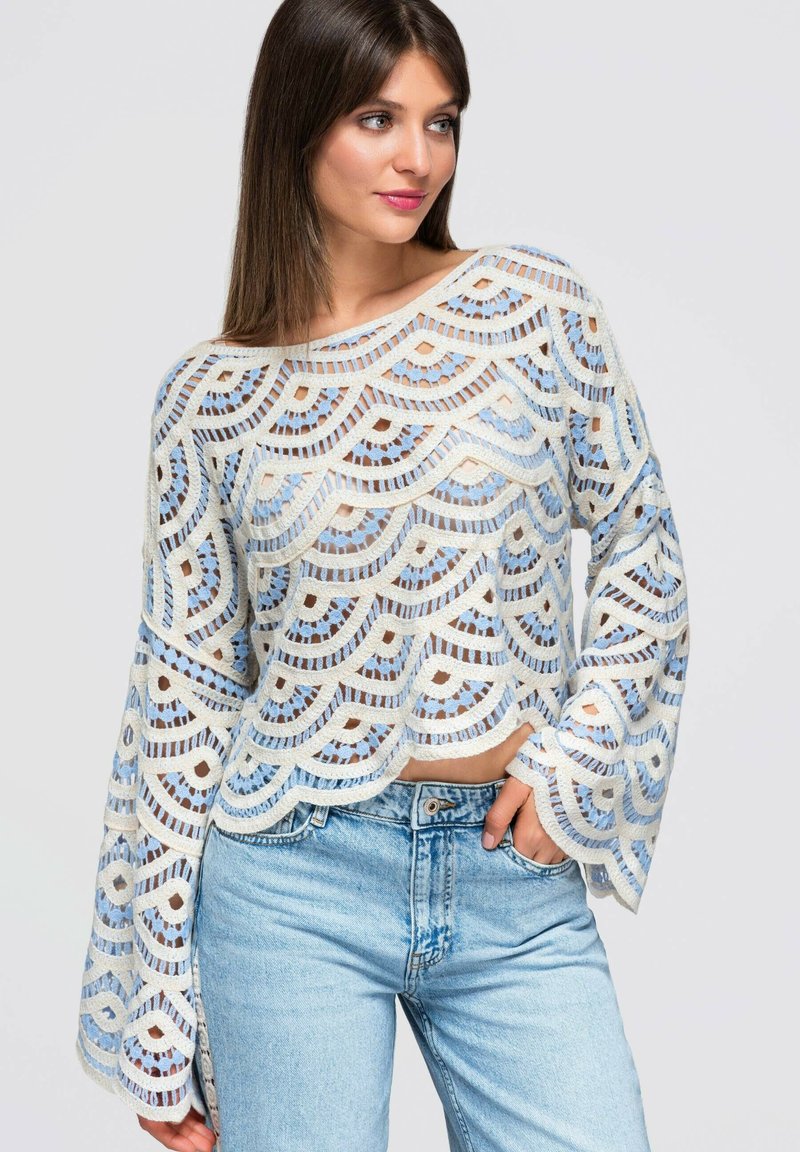 Femme portant un haut en crochet blanc et bleu à manches longues avec des bords en feston et un jean bleu clair, posant avec une main dans la poche.