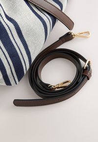 Sac en tissu rayé avec une bandoulière en cuir marron équipée de fermoirs dorés, enroulée à côté du sac sur une surface claire.