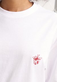 Tricou alb din bumbac, cu un mic design brodat roșu reprezentând o cutie de mâncare la pachet cu bețișoare, situat în zona stângă a pieptului.