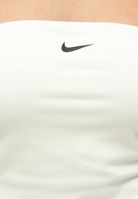 Nahaufnahme einer Person, die ein weißes trägerloses Oberteil trägt, auf dem mittig an der Brust ein schwarzes Nike-Swoosh-Logo abgebildet ist.