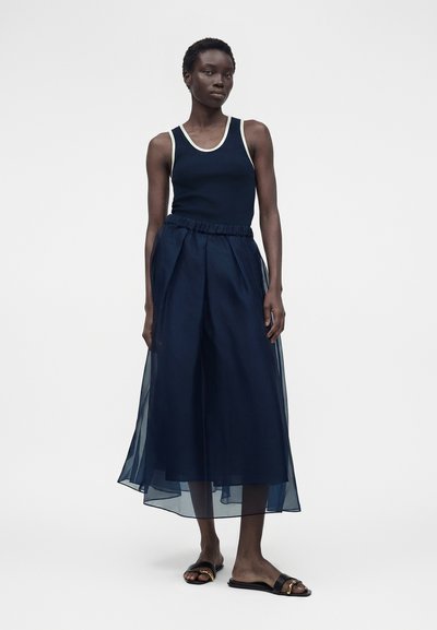 Γυναίκα που στέκεται φορώντας μπλε σκούρο αμάνικο τοπ με λευκή μπορντούρα και μπλε σκούρο φαρδιά διάφανα παντελόνια με μαύρα σανδάλια slide.