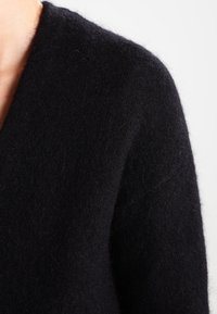 Černý pletený cardigan s hlubokým V-výstřihem, měkkou texturou a mírně chlupatým povrchem, který zdůrazňuje útulnost materiálu oděvu.