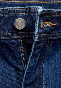 Vue en plongée d'un jean en denim présentant une fermeture à pression métallique et une fermeture éclair, avec un tissu bleu marine, des coutures visibles et des bords effilochés près du bouton.