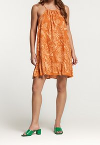 Woman wearing an orange floral halter dress with ruffle hem and green heeled open-toe mules, standing against a plain background.