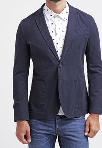 Marinblå kavaj gjord av lättviktig tyg, med två frampocket, en enkel knappstängning och en skarp krage. Buren över en vit skjorta med prickar.