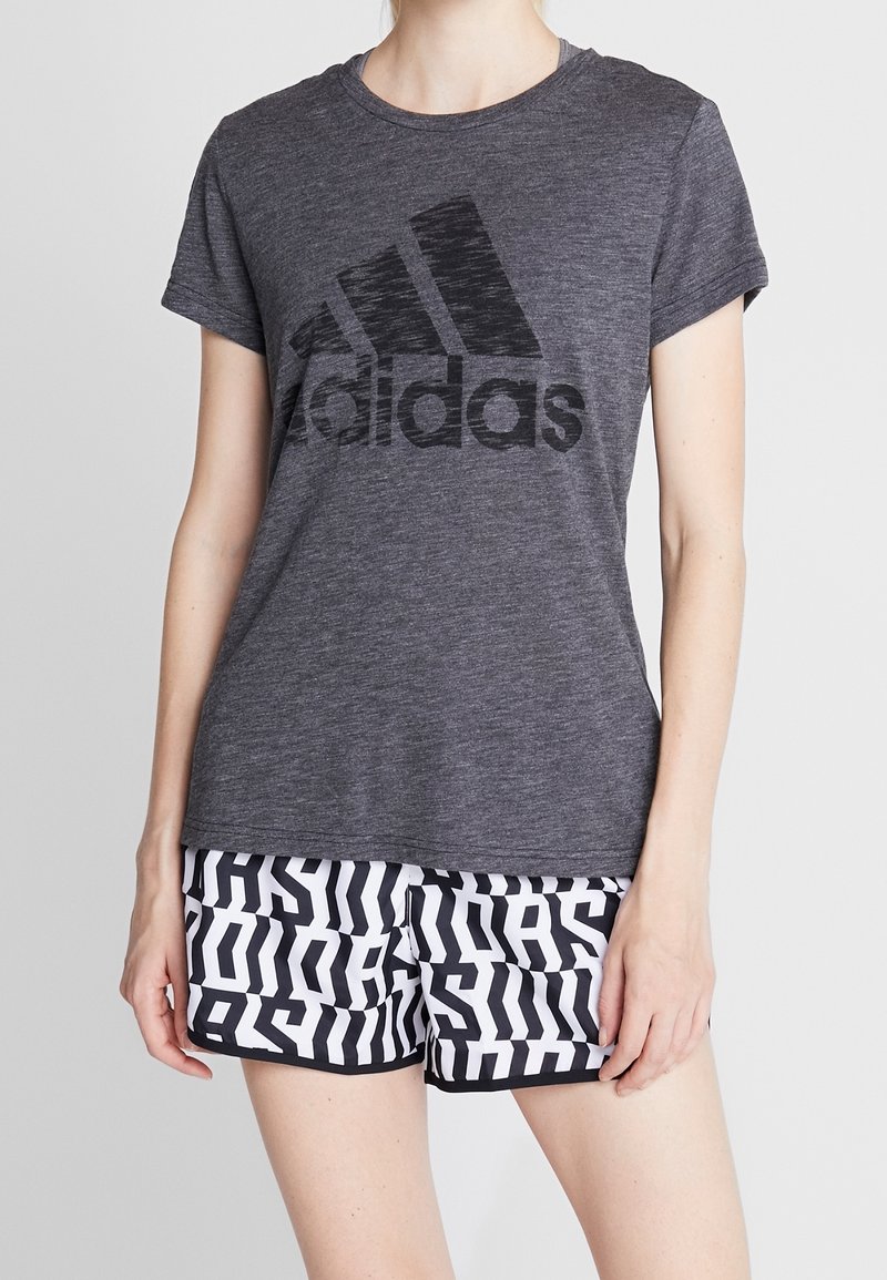 Grå t-shirt med korte ærmer og et stort svart Adidas-logo, kombineret med mønstrede sorte og hvide shorts, der viser gentaget branding.