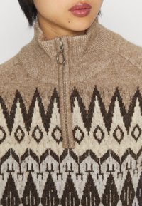 Pullover lavorato a maglia con colletto alto, dotato di zip. Motivo geometrico marrone e crema su uno sfondo beige caldo e texturizzato.