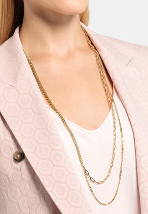 Collier en métal doré à plusieurs épaisseurs, présentant des styles de maillons de chaîne alternés, porté sur un haut rose clair, complété par un blazer à motifs.