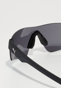Lunettes de soleil de sport noires avec un design enveloppant, verres teintés gris et un logo Puma argenté évident sur le côté.