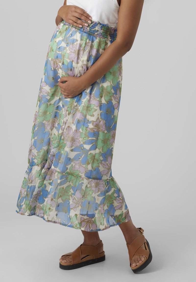 Personne enceinte portant un haut blanc, une jupe à motif floral avec des fleurs bleues et vertes, et des sandales compensées marron, les mains reposant sur le ventre.