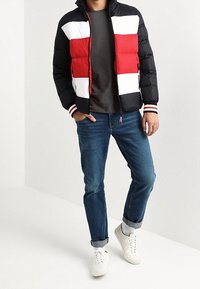 Man som bär en svart, röd och vit färgblockerad pufferjacka, mörkgrå t-shirt, uppvikta blå jeans, vita strumpor och vita sneakers.