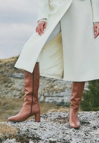 Bocanci din piele maro, până la genunchi, cu toc pătrat, textură netedă și fermoar lateral, purtați cu o haină albă și stând pe un teren stâncos.