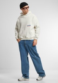 Gray hooded sweatshirt with front pocket and blue logo, paired with loose blue jeans and silver sneakers with reflective accents. Black beanie and sunglasses.