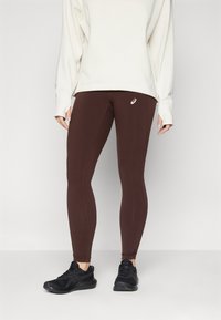 Leggings atléticos marrones hechos de material elástico, con un diseño ajustado y costuras laterales. Combinados con un sudadera de corte holgado en crema y zapatillas negras.