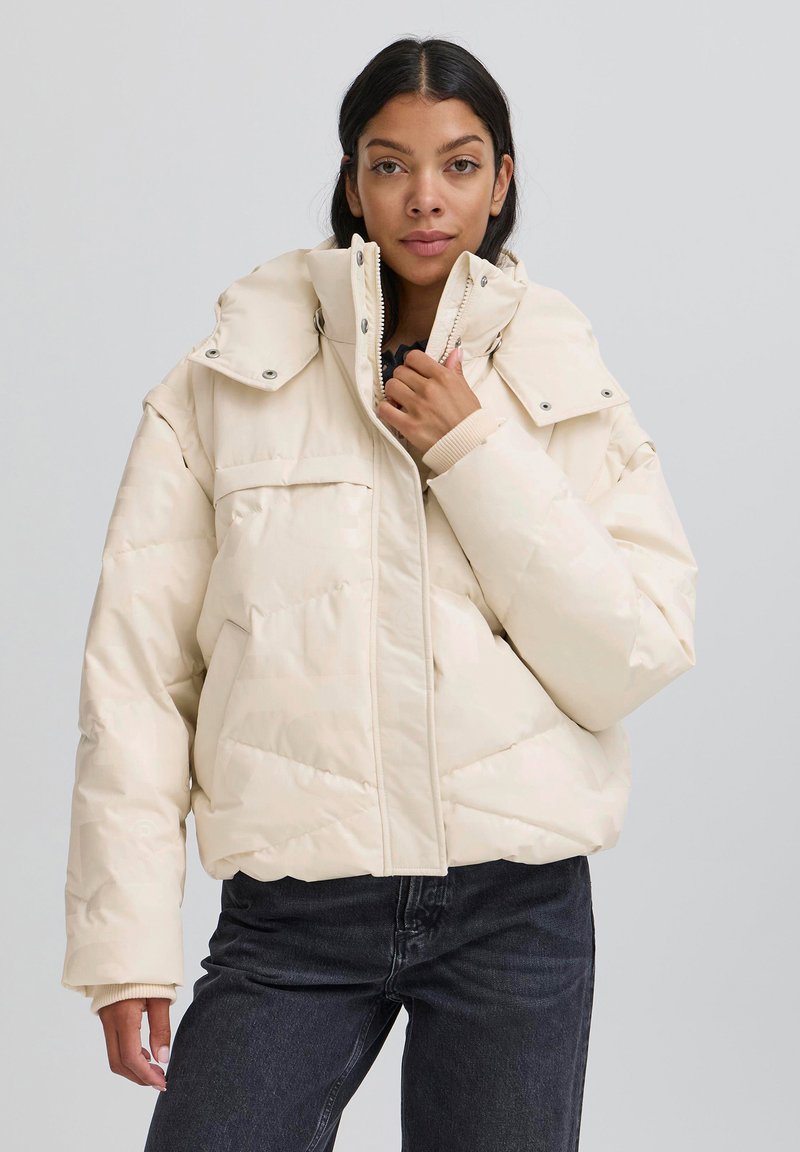 Vieglā bēša krāsas pufi jaka ar augstu apkakli, divām priekšējām kabatām, dekoratīvu šūšanas rakstu un elastīgām aprocēm. Rāvējslēdzēja aizdare ar aizdares pogām.