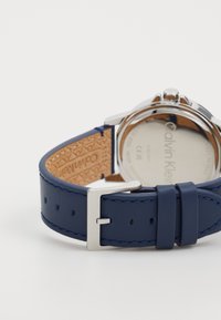 Montre Calvin Klein avec un bracelet en cuir bleu, dont la quincaillerie est en argent, des coutures contrastées, et un fond en acier inoxydable.
