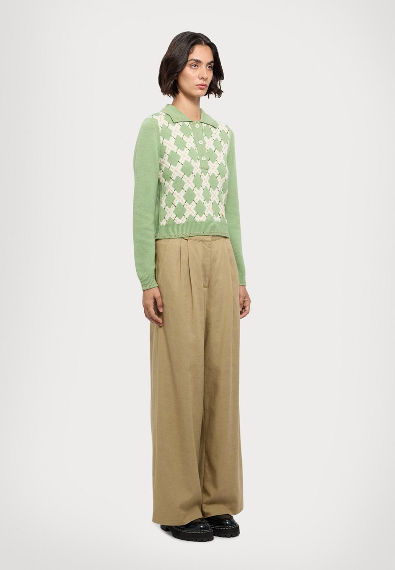 Maglione verde lavorato a maglia con motivo a diamante, colletto rotondo e bottoni frontali, abbinato a pantaloni beige a gamba larga e scarpe platform nere.