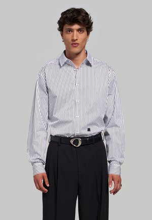 Jeune homme portant une chemise blanche à fines rayures verticales noires et un pantalon noir avec une ceinture noire et une boucle argentée, debout devant un fond uni.