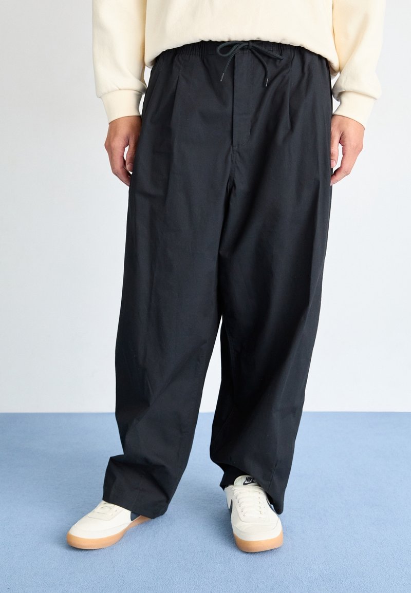 Svart, löst sittande byxor med elastisk midja, sidfickor och dragsnöre. Stylad med vita sneakers på en blå yta.