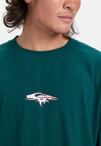 T-shirt en coton bleu canard avec un col rond. Dispose d'un logo stylisé blanc et rouge sur la poitrine. Texture lisse avec une coupe décontractée.